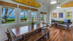 Uma área de refeições com mesa de madeira e bancos, janelas amplas com vista para o oceano e pranchas de surf fixadas no teto.