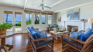 Uma sala de estar ampla com móveis de vime, lareira e grandes janelas com vista para o mar.