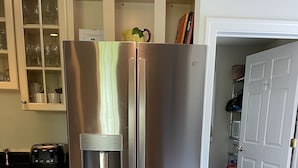 A stainless steel refrigerator with a water dispenser, a microwave, and a toaster. There are white cabinets with glass doors and a tiled floor.