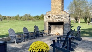 A stone fireplace with Adirondack chairs around it, a concrete patio, and a grassy field in the background.