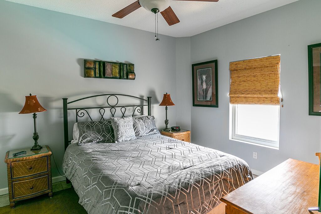 Quarto com cama, duas mesas de cabeceira, ventoinha no teto, janela com persianas e obras de arte nas paredes.