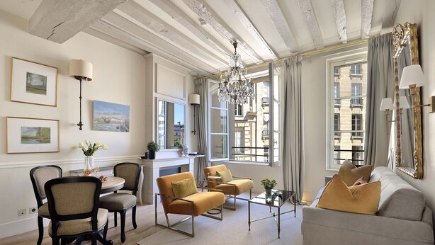 Un salon bien éclairé, avec un canapé, des fauteuils, une table basse et une cheminée. Des tableaux encadrés ornent le mur, et la fenêtre offre une vue sur les bâtiments environnants.