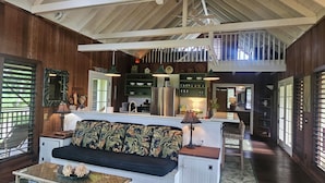 A spacious living area with a high ceiling, wooden beams, a central island with seating, a kitchen area, and a staircase leading to an upper level. - Beautiful Plumeria Cottage-Anini Beach Plantation Style Home  (Anini Beach)
