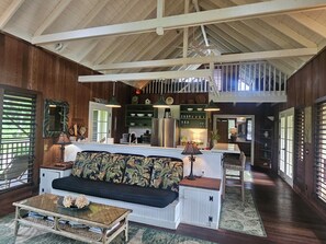 Amplia sala de estar con techos altos, vigas de madera, una isla central con asientos, zona de cocina y una escalera que conduce a un nivel superior.