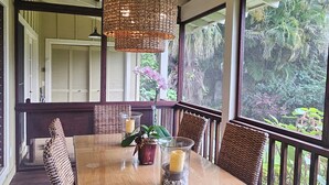 A wooden dining table with wicker chairs, a hanging wicker light fixture, and a view of greenery through large windows. - Beautiful Plumeria Cottage-Anini Beach Plantation Style Home  (Anini Beach)