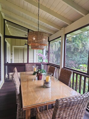 Una mesa de comedor de madera con sillas de mimbre, una lámpara colgante de mimbre y vistas a la vegetación a través de amplias ventanas.