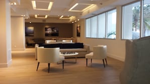 A modern living room with a sofa, two armchairs, and a coffee table. There is a large window on the right side, and a television is mounted on the wall. - SOBE ~ ART DECO DISTRICT ~ SOUTH BEACH ~ RITZ CARLTON       Condo on the Water (Miami Beach)