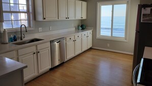 Una cocina con gabinetes blancos, lavavajillas de acero inoxidable y vista al océano a través de un ventanal grande.