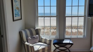 Una habitación con un ventanal grande que ofrece vistas a la playa, un sillón a rayas, un taburete pequeño y un televisor sobre un mueble bajo.