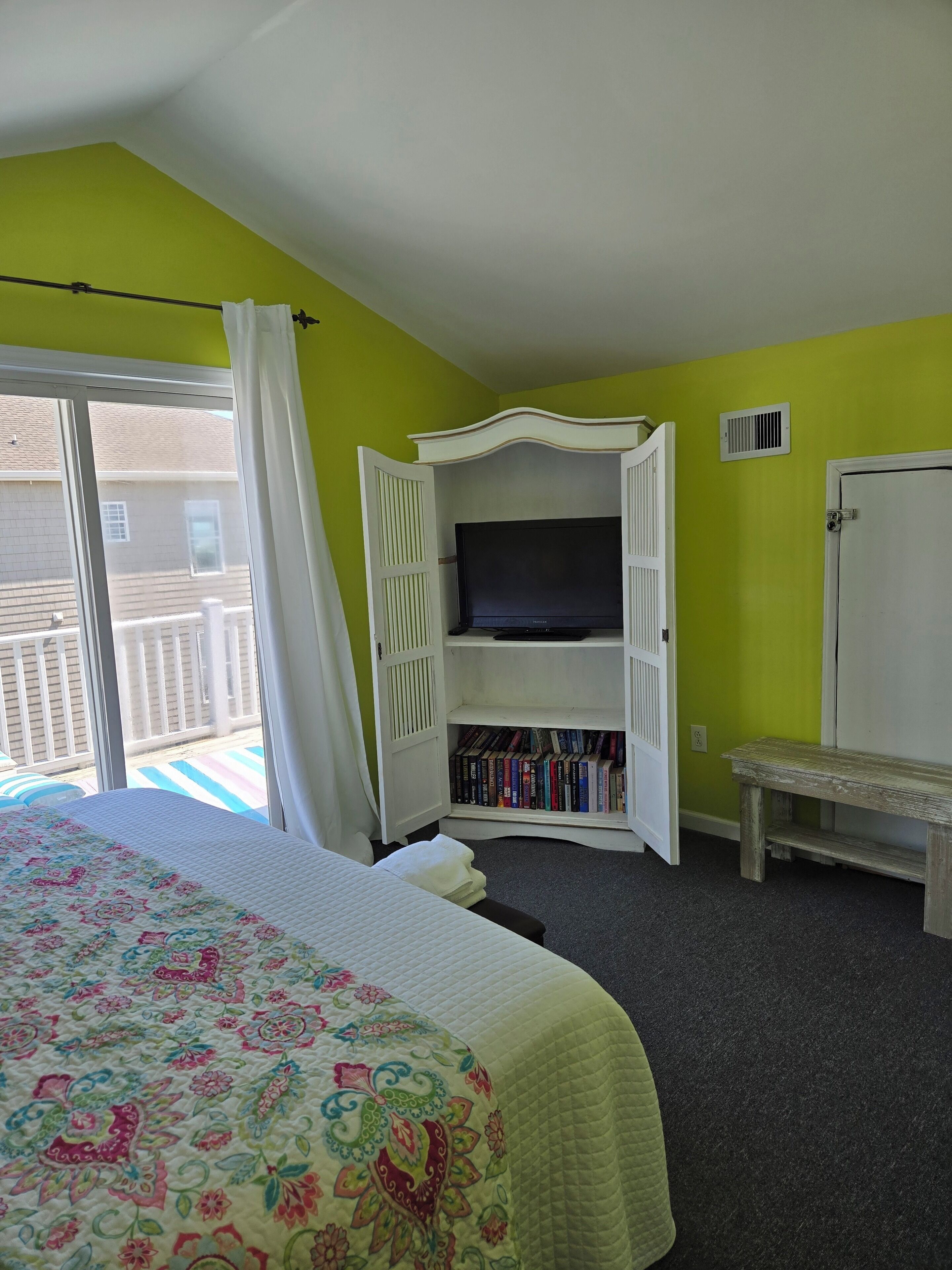 A hotel room with a bed, a television, a bookshelf, and a bench.
