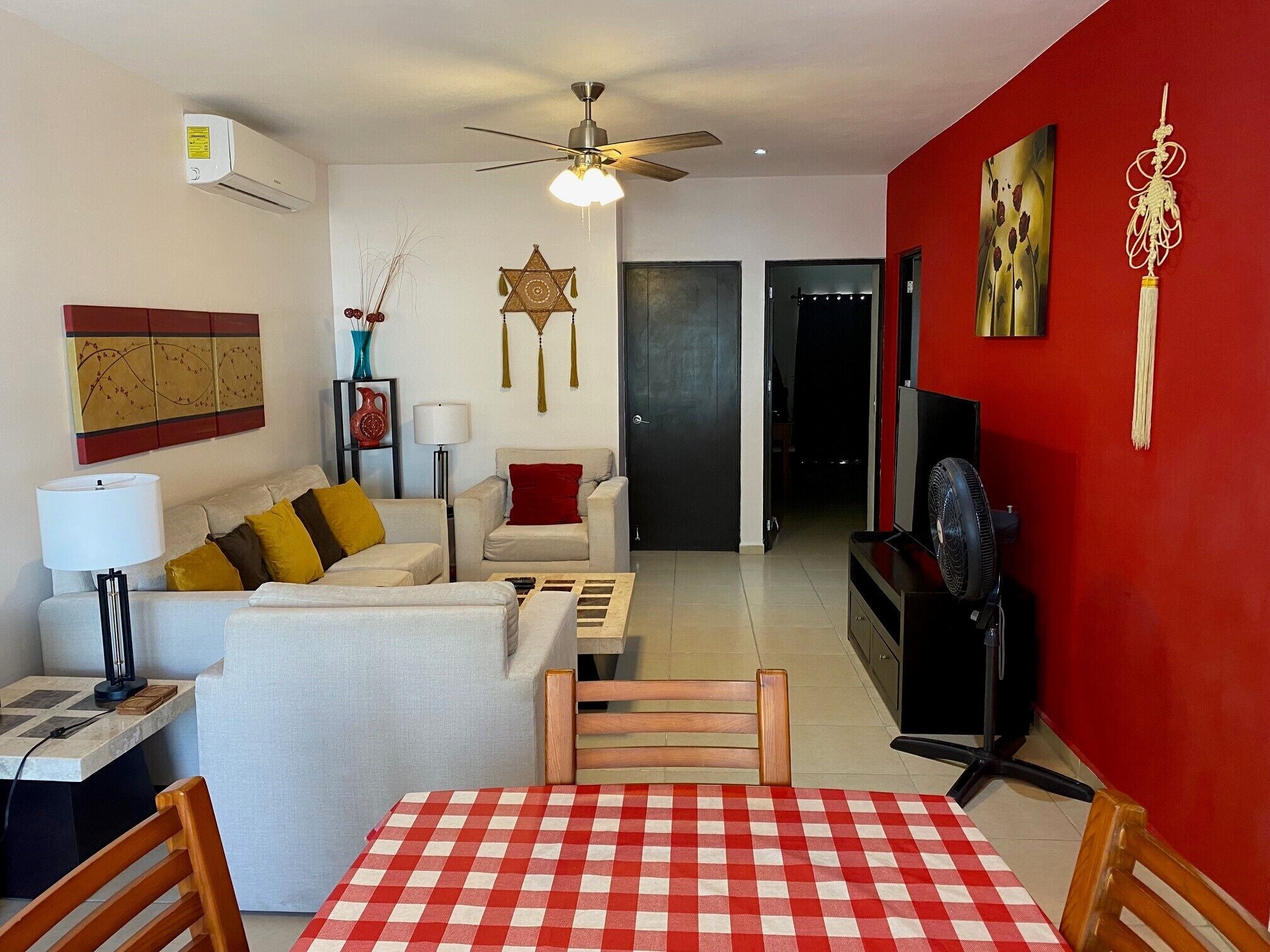 Un salon avec un mur rouge, un ventilateur de plafond, un canapé blanc agrémenté de coussins jaunes, une table à carreaux rouges et blancs, et un meuble pour la télévision.