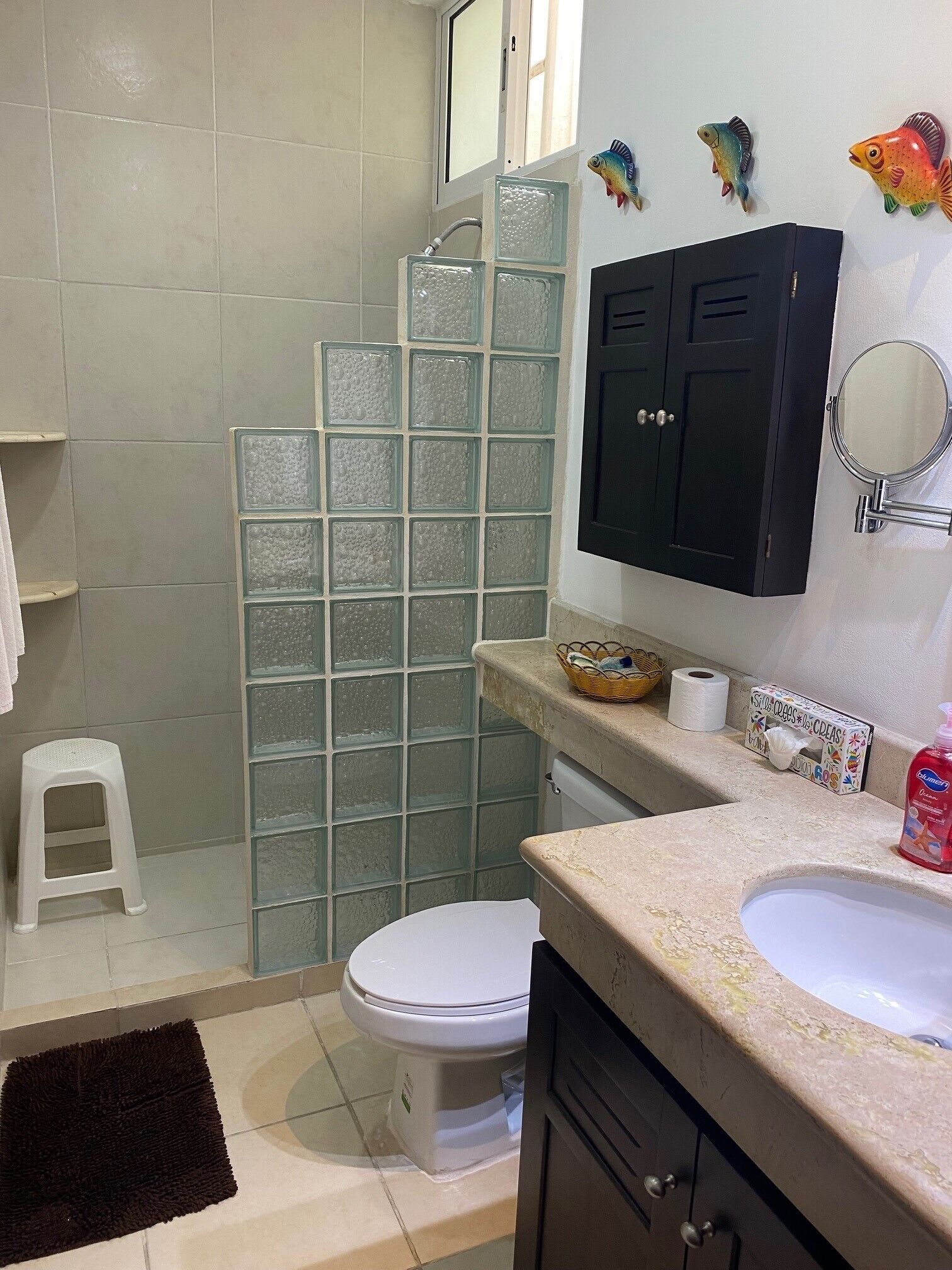 Een badkamer met een toilet, een wastafel, een glazen douchecabine, een klein krukje en decoratieve vissen aan de muur.