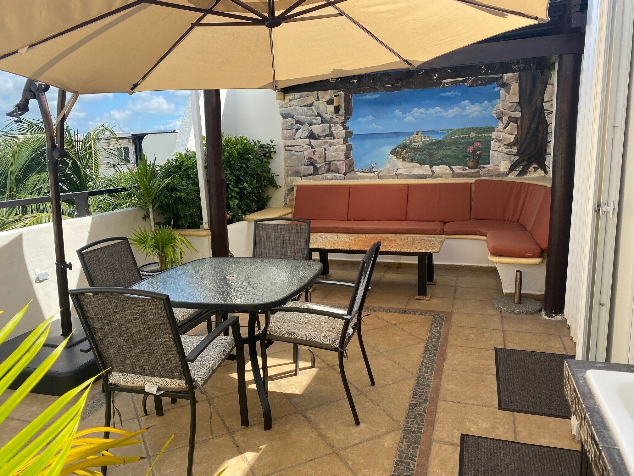An outdoor patio with a table and chairs, a cushioned bench, and a view of a coastal landscape through a stone wall.