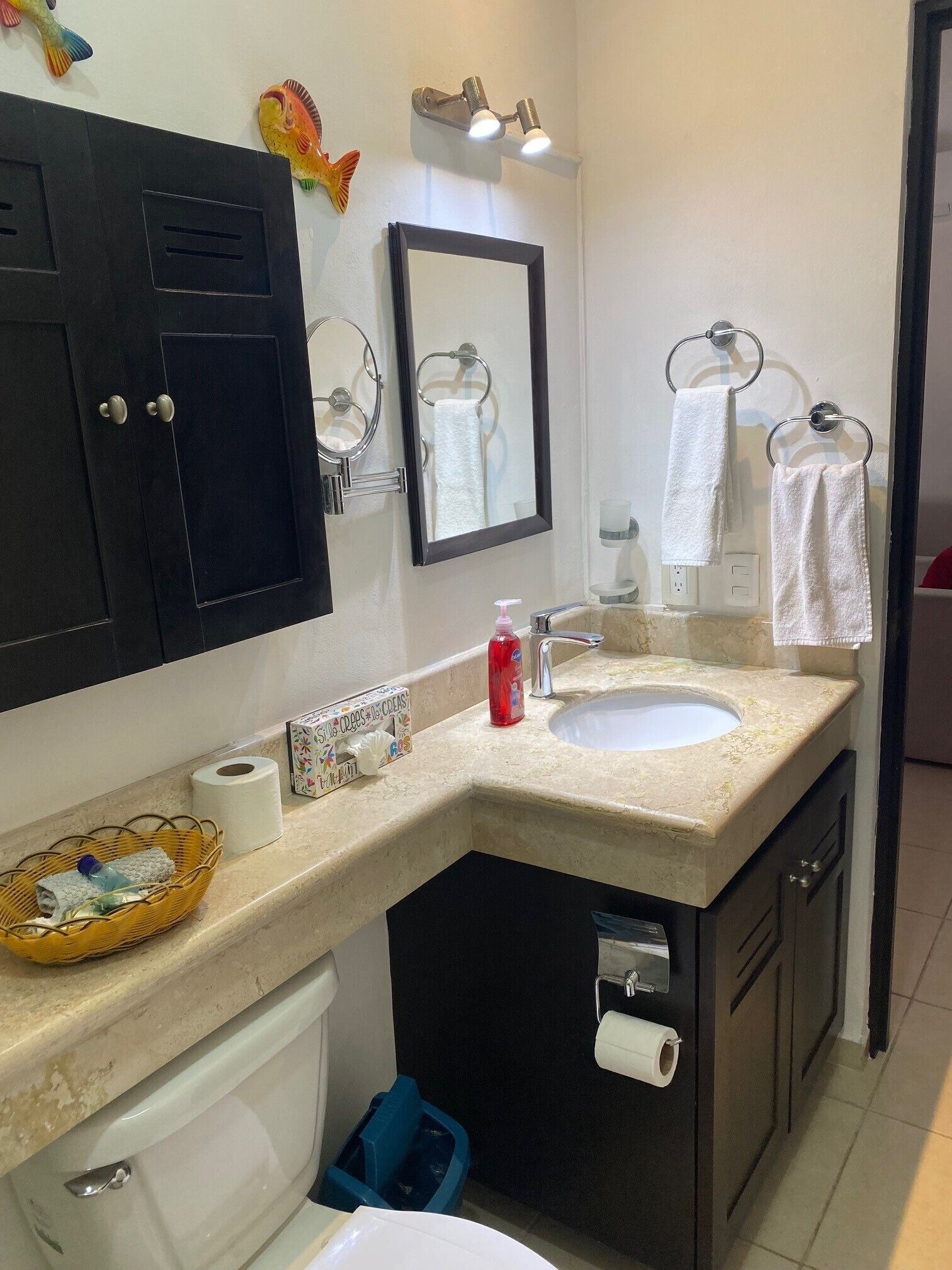 A bathroom with a double sink, a mirror, a towel rack, a basket with toiletries, and a trash bin.