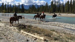 Un grupo de personas cabalgando a lo largo de la orilla de un río, con un bosque y montañas al fondo.