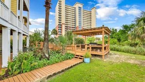 A hotel with a wooden deck, lush greenery, and a view of high-rise buildings in the background. - 3BR/3BA - Beach, pool, lagoon front and MORE!  (Gulf Shores)