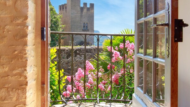 Porta aperta che dà su un balcone con vista su un castello, circondato da fiori rosa e vegetazione.
