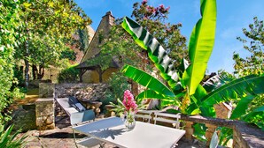 Patio esterno con tavolo e sedie in pietra, circondato da vegetazione lussureggiante e un edificio in pietra con tetto di paglia.