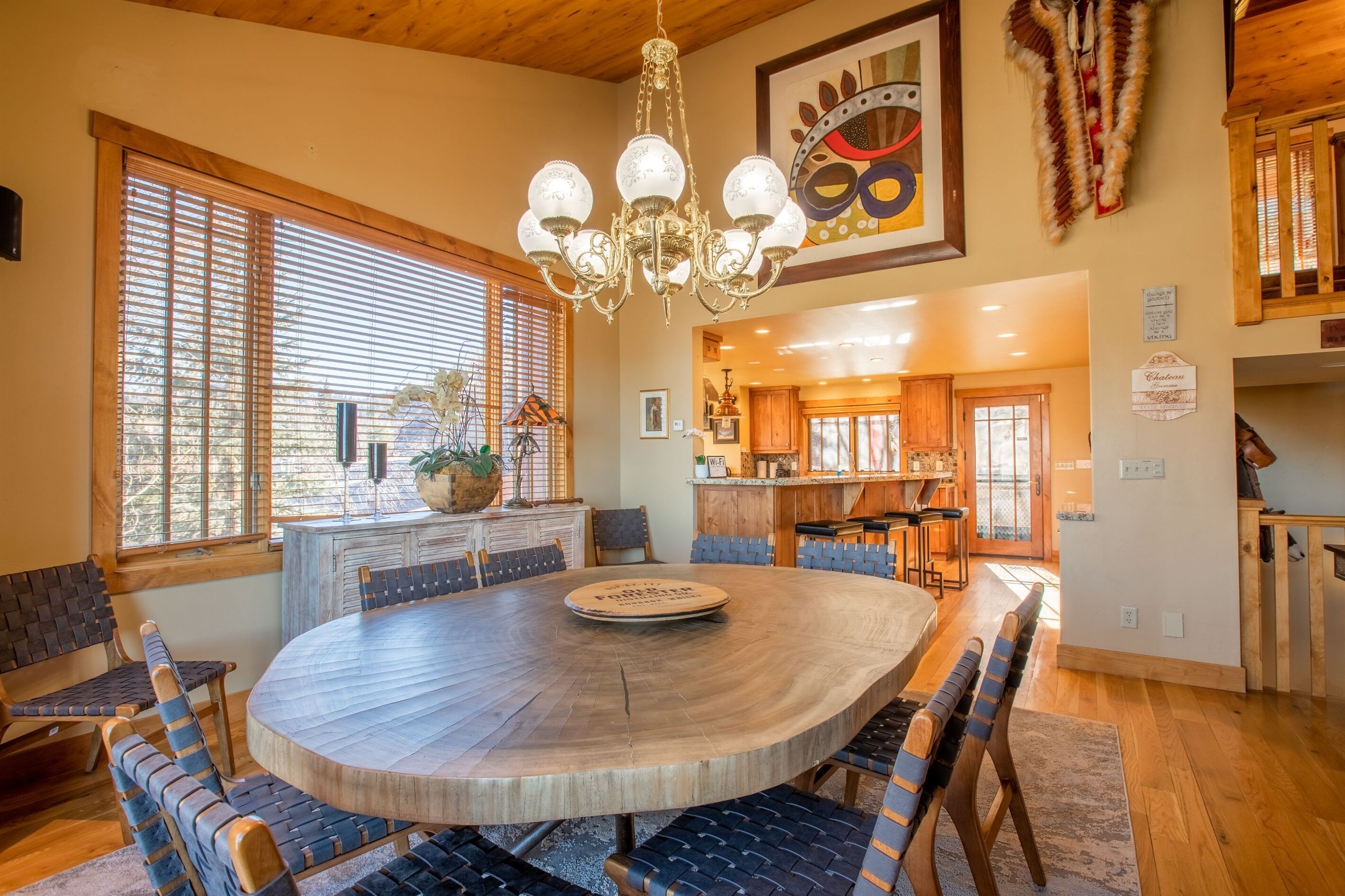 A spacious dining area with a round wooden table and chairs, a chandelier, and large windows with blinds.