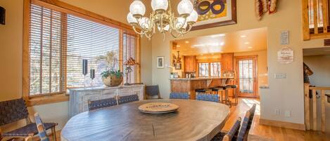 A spacious dining area with a round wooden table and chairs, a chandelier, and large windows with blinds.