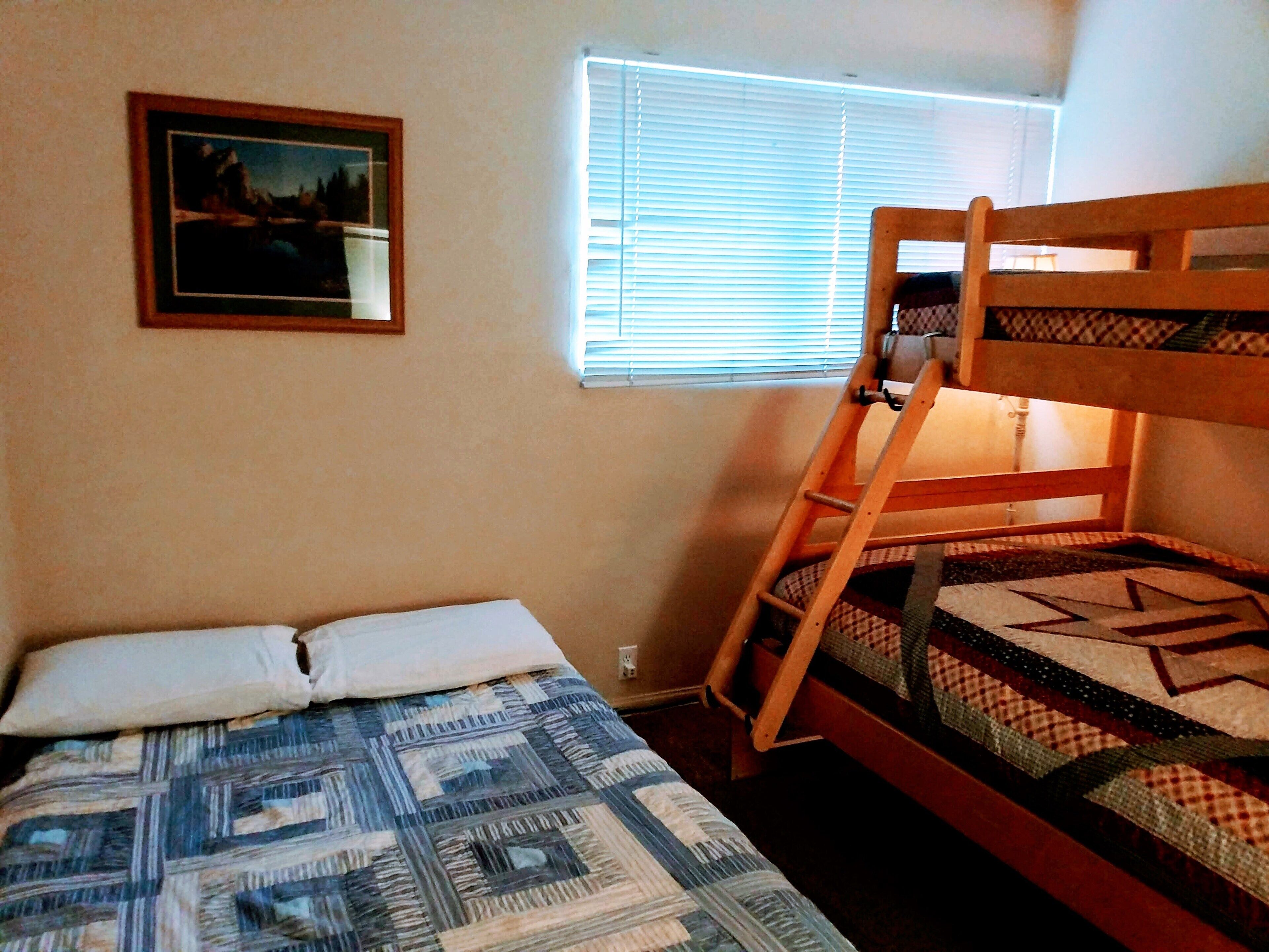 Ein Etagenbett mit einer Leiter, ein Einzelbett mit einem gemusterten Bettbezug, ein Fenster mit Jalousien und ein gerahmtes Bild an der Wand.