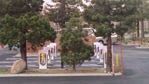 A parking area with electric vehicle charging stations, trees, and a building in the background.