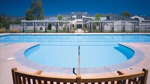 Una piscina con muelle de madera en primer plano, un porche cubierto al fondo y un cielo azul despejado.