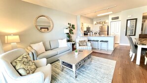 A modern living room with a sofa, coffee table, and a dining area with a kitchen in the background. - Happy Hours@Shores of Panama | Beachfront Resort Pools, Hot Tub & Tiki Bar (Panama City Beach)