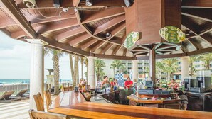 A beachside bar with wooden tables and chairs, a view of the ocean, and palm trees. - Happy Hours@Shores of Panama | Beachfront Resort Pools, Hot Tub & Tiki Bar (Panama City Beach)