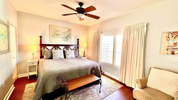 Une chambre à coucher comprenant un lit, des tables de chevet, un banc, une chaise, un ventilateur de plafond et une fenêtre avec des rideaux.