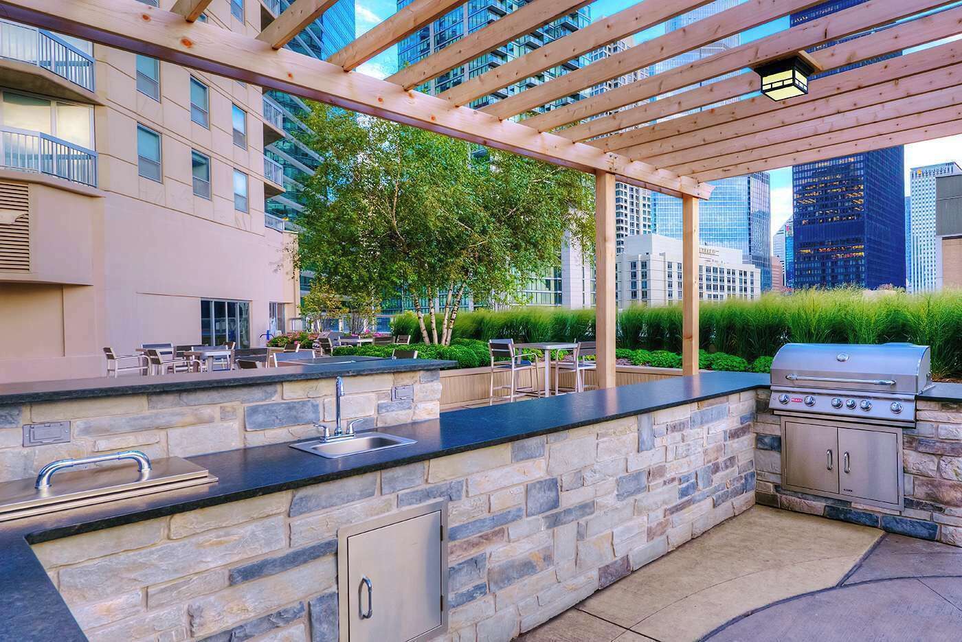 Et udendørs køkken med en bordplade af sten, en vask og en grill, omgivet af en pergola og grønne planter.