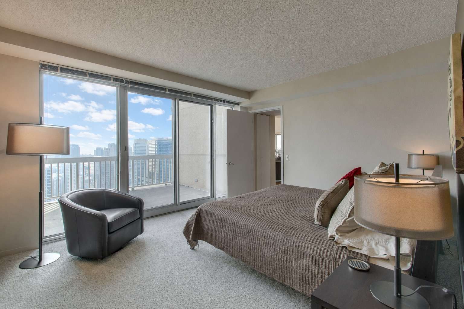 A hotel room with a large window, a bed, a chair, and a desk with a lamp.