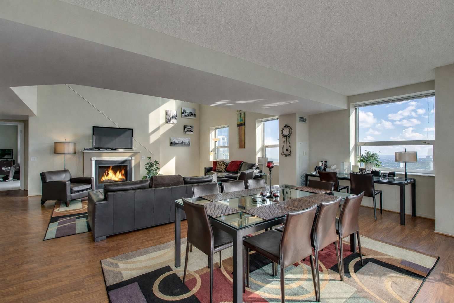 A modern living room with a fireplace, a dining area with a glass table, and a large window offering a city view.