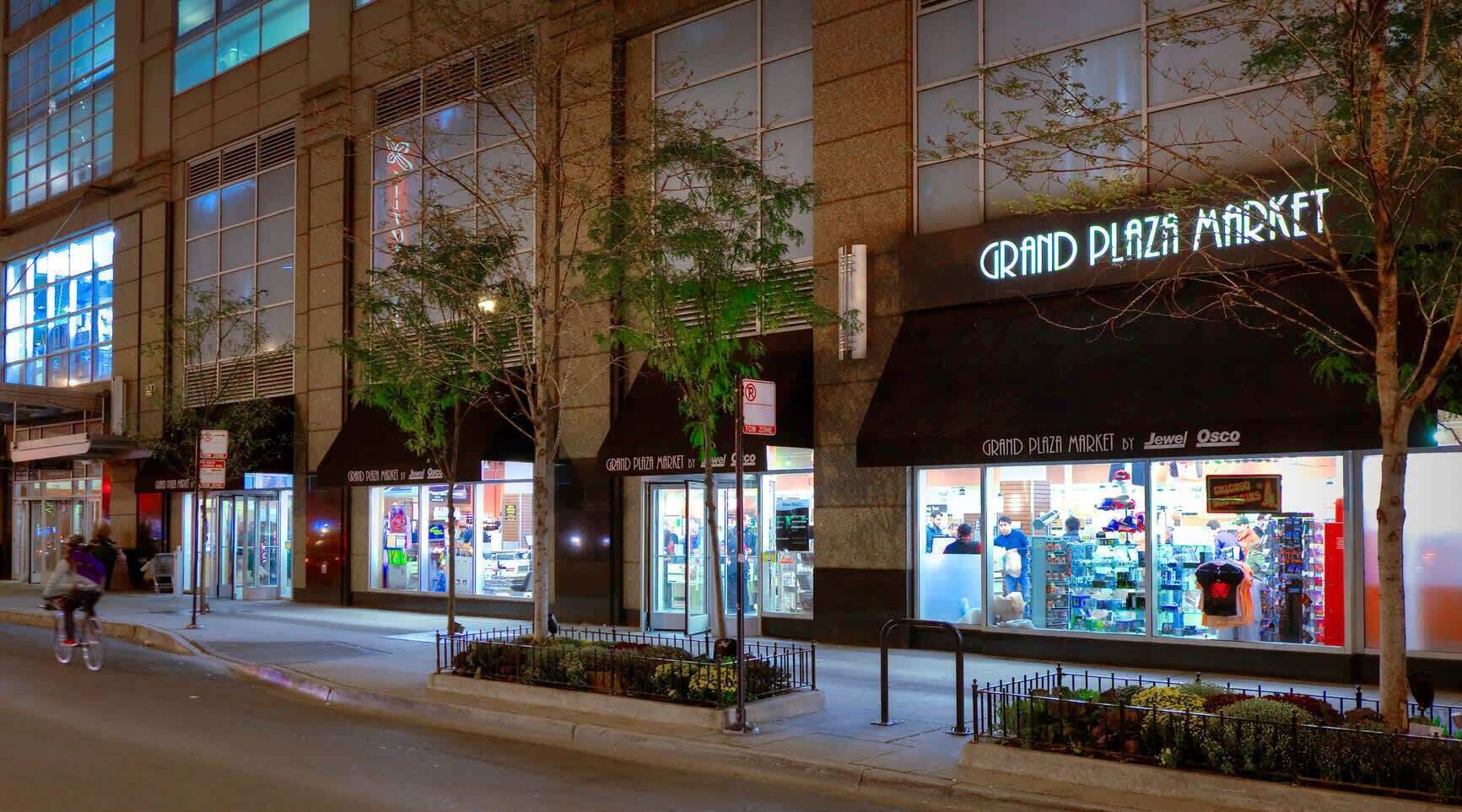 A Grand Plaza Market with multiple storefronts, including Jewel and Osco, on a city street at night.