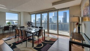 A modern living room with a glass dining table, comfortable seating, a large window with a city view, and a balcony with a railing. - 2-Story Penthouse Views, Fireplace, Pool, Gym (Chicago)