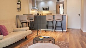 Een moderne woonkamer met een bank, twee barkrukken, een salontafel en een keukenhoek met een magnetron en een koelkast.