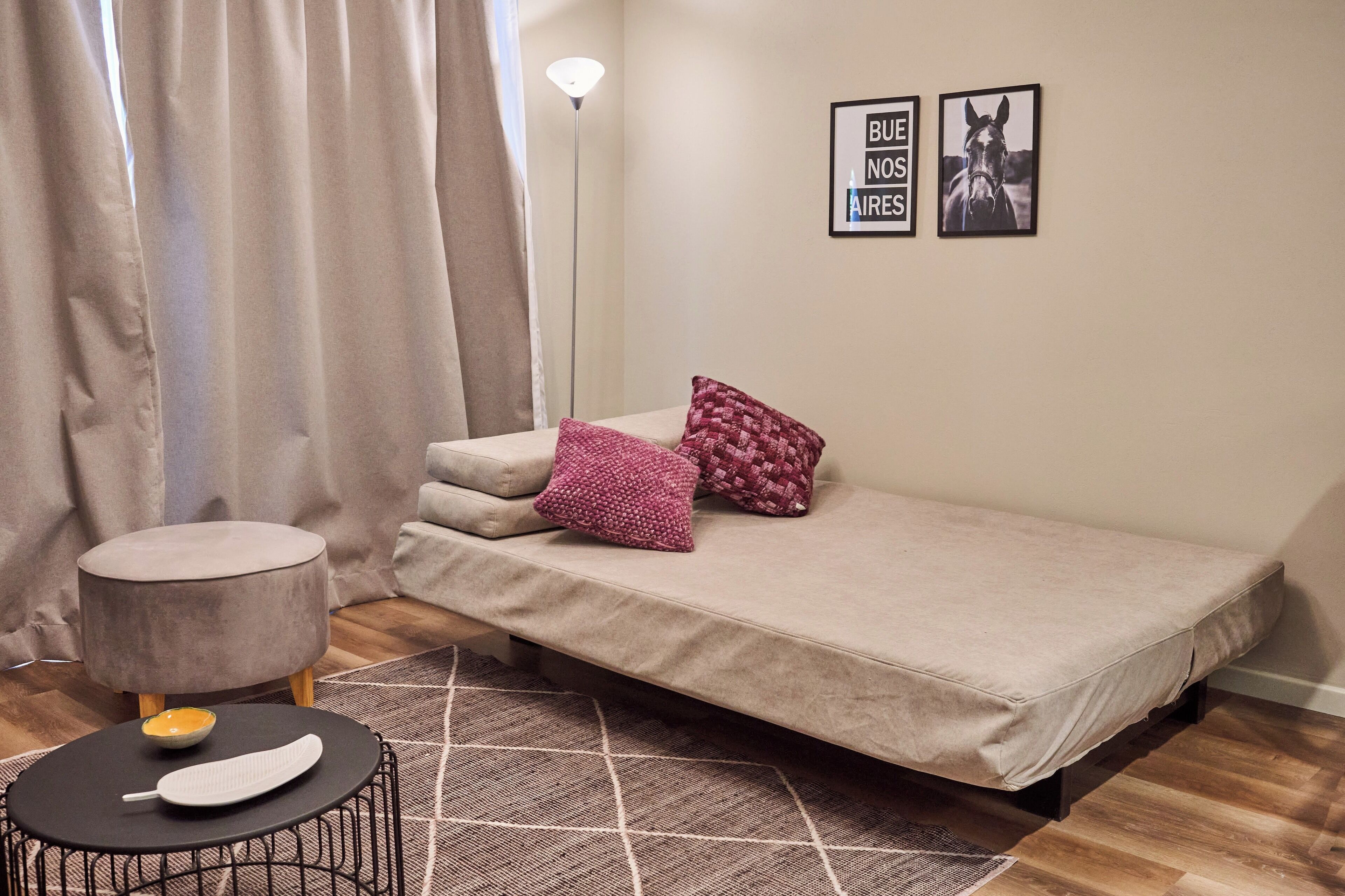 A small, cozy bedroom with a single bed, a round ottoman, a small table with a candle and a feather, and a geometric-patterned rug.