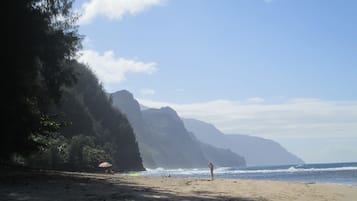 A beach with a person standing, a colorful umbrella, and a mountain in the background.