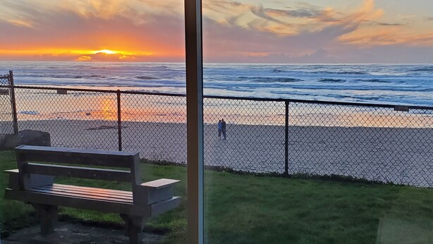 Vista del océano al atardecer a través de un ventanal, con un banco y una valla en primer plano.