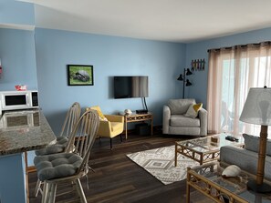 A living room with a blue wall, a flat-screen TV, a sofa, a chair, a coffee table, and a lamp. - Lovely Oceanfront Condo w/ Vast Top Floor Views; Pool; WiFi/Cable; Quiet; 2 Max (Carolina Beach)