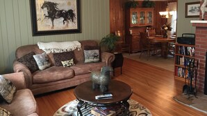 A cozy living room with a brown leather sofa, a coffee table with books and a remote, a framed horse painting, and a dining area with wooden furniture.