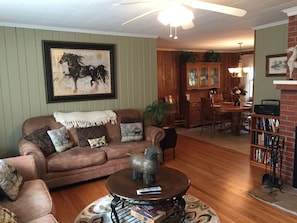 A cozy living room with a brown leather sofa, a coffee table with books and a remote, a framed horse painting, and a dining area with wooden furniture.