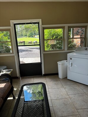 A room with a door leading to a grassy area, a washing machine, a trash bin, and a table with a woven table runner.