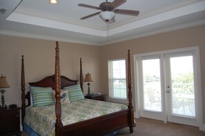 A four-poster bed with a floral bedspread, two bedside lamps, and a ceiling fan in a bedroom with beige walls and a large window with blinds.