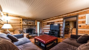 A log cabin interior with a fireplace, a television, and a cozy seating area with sofas and a red ottoman. - Stunning Family/Friends Retreat,110 Private Acres, Renovated Historic Home (Madison)