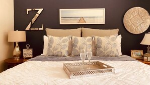 A neatly made bed with a tray and two glasses, a large 'Z' decoration on the wall, a framed picture, and a round textured wall piece.