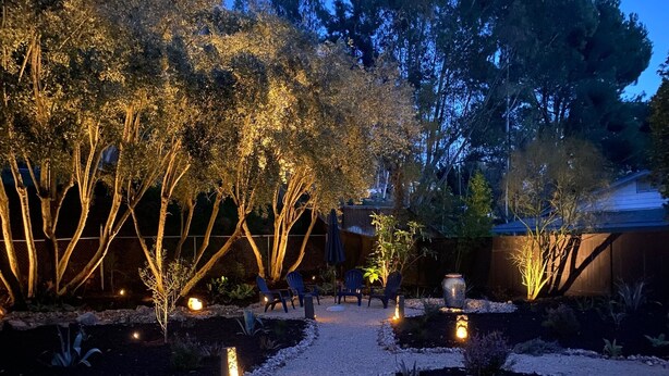 A garden pathway with trees and chairs, surrounded by a wooden fence and illuminated by ground lights.