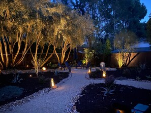 Caminho no jardim com árvores e cadeiras, cercado por uma vedação de madeira e iluminado por luzes no chão.