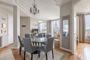 A dining area with a round table, grey upholstered chairs, a chandelier, and a view of the Eiffel Tower. - Stunning & Bright, Sleeps 6, w/Big Balcony and Amazing Paris Views (Paris)