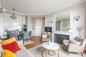 A modern living room with a sofa, chairs, a coffee table, a fireplace, and a kitchen area with cabinets and appliances. - Stunning & Bright, Sleeps 6, w/Big Balcony and Amazing Paris Views (Paris)